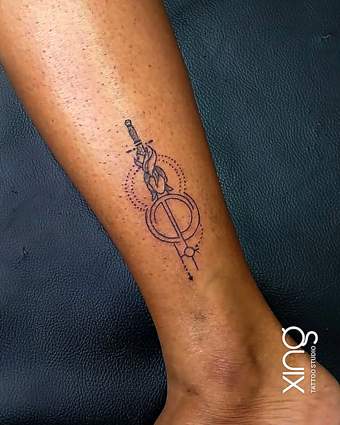 Fine-line sword and circle emblem tattoo on ankle